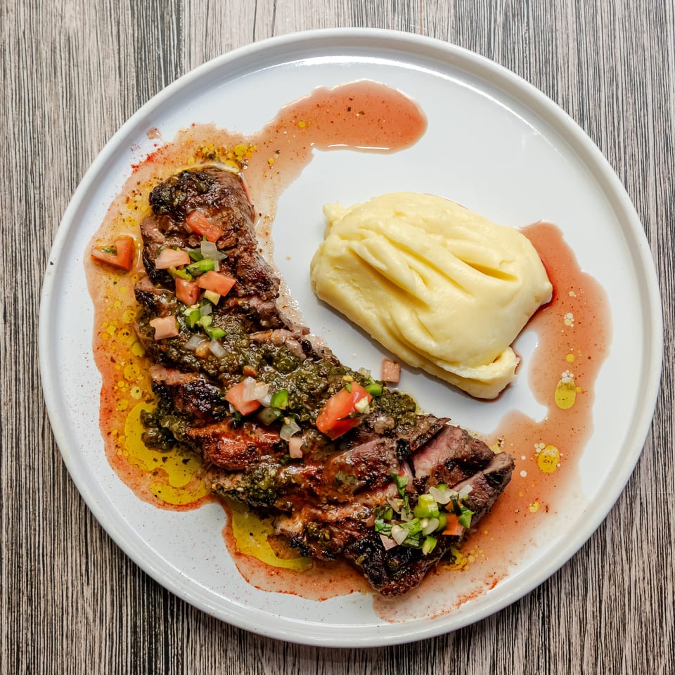 Grilled sliced steak topped with chimichurri and diced vegetables, served with mashed potatoes and colorful sauces on a white plate.