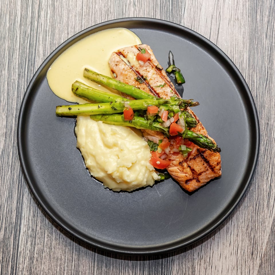 Grilled salmon with asparagus, mashed potatoes, creamy sauce, and diced tomato relish on a black plate.