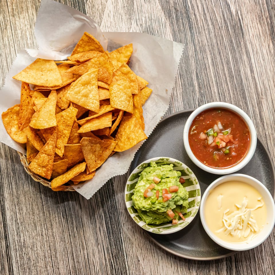 Basket of tortilla chips served with salsa, guacamole, and creamy cheese dip on a wooden table.