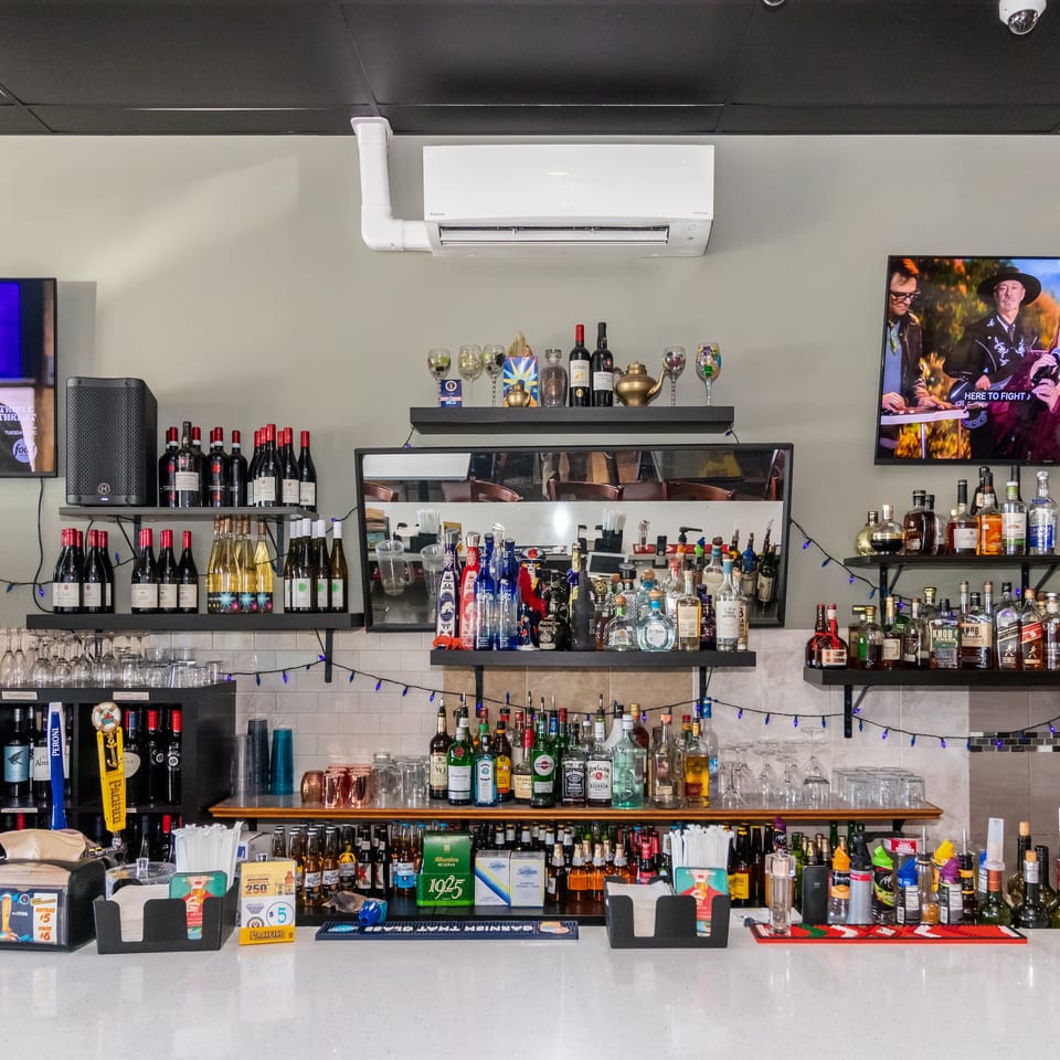 Well-stocked restaurant bar with liquor shelves, wine bottles, televisions, glassware, and colorful string lights.